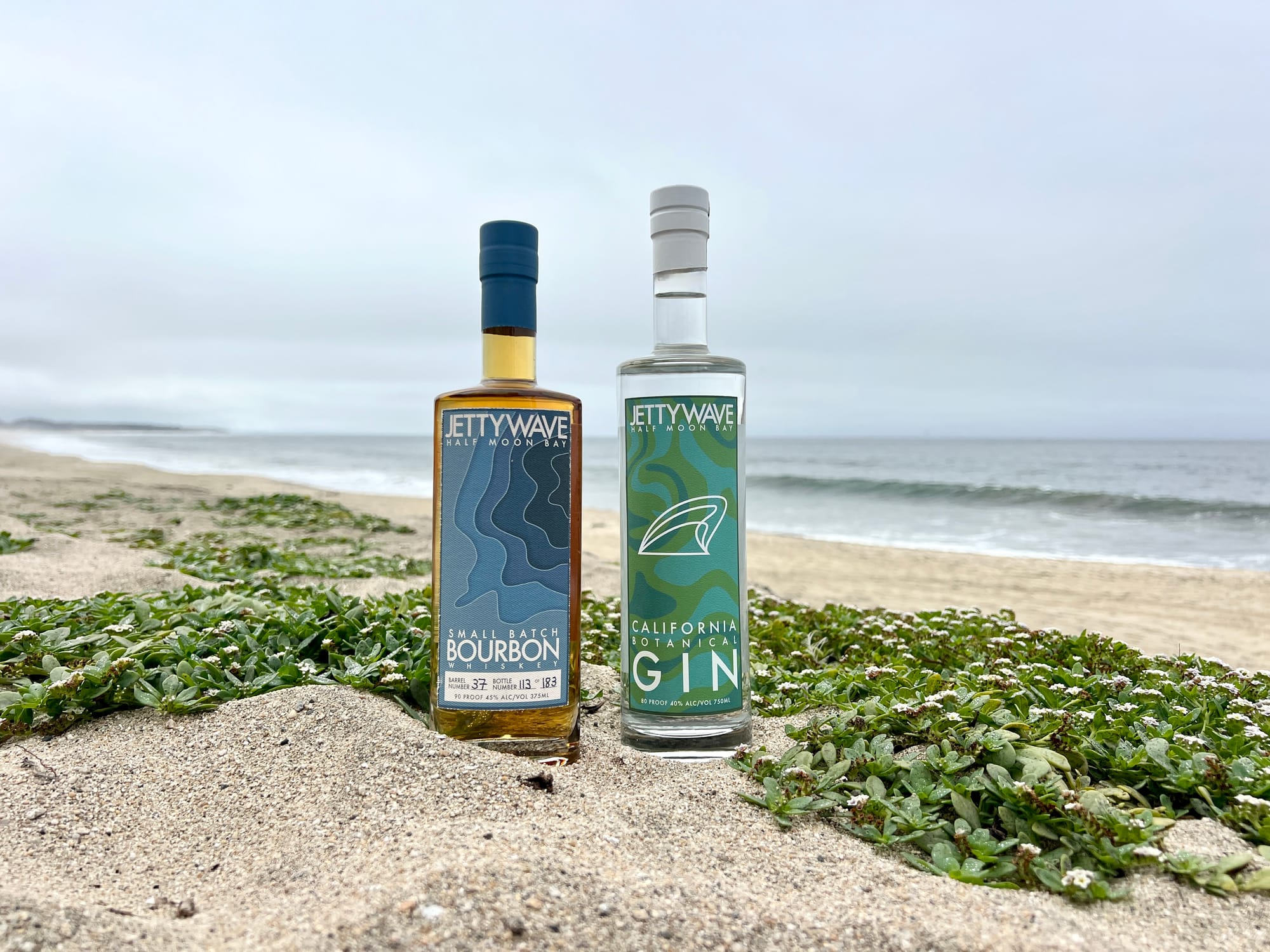 A bottle of Jettywave Small Batch Bourbon and California Botanical Gin on a sand dune overlooking Half Moon Bay.