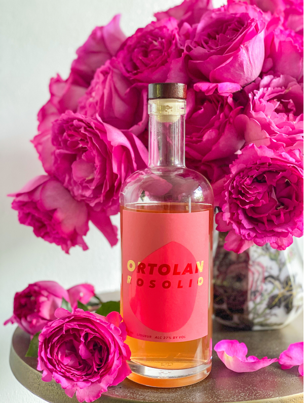 A bottle of Ortolan Rosolio sits atop a cafe table surrounded by pink rose blossoms.