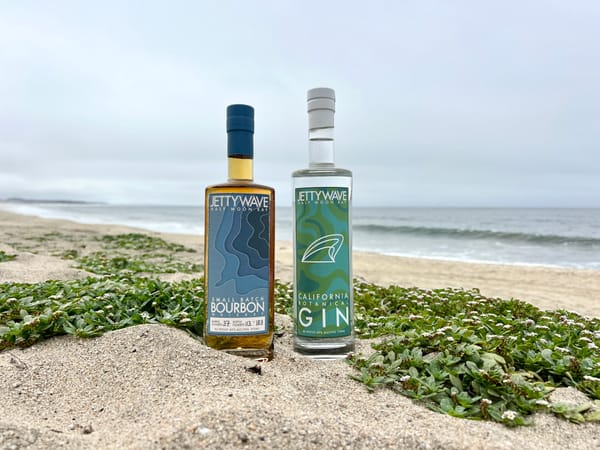 A bottle of Jettywave Small Batch Bourbon and California Botanical Gin on a sand dune overlooking Half Moon Bay.